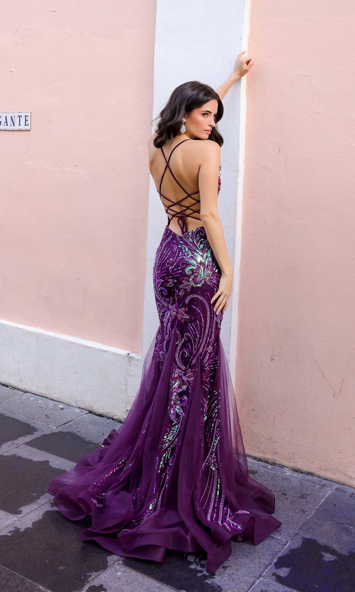 A woman in the Nox Anabel Tight Sequin Mermaid Prom Dress C1416 with a strappy open back poses against a pale pink wall, looking over her shoulder.